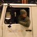 A person wearing U.S. Air Force fleece and an olive green beanie is seated in the cab of a Woodward Aviation Services ground support vehicle inside what appears to be an aircraft hangar. The individual is looking to the side, suggesting they are maneuvering or monitoring their surroundings while operating the vehicle. The vehicle's door panel displays Woodward Aviation Services contact information including phone, fax, and Unicom frequency.