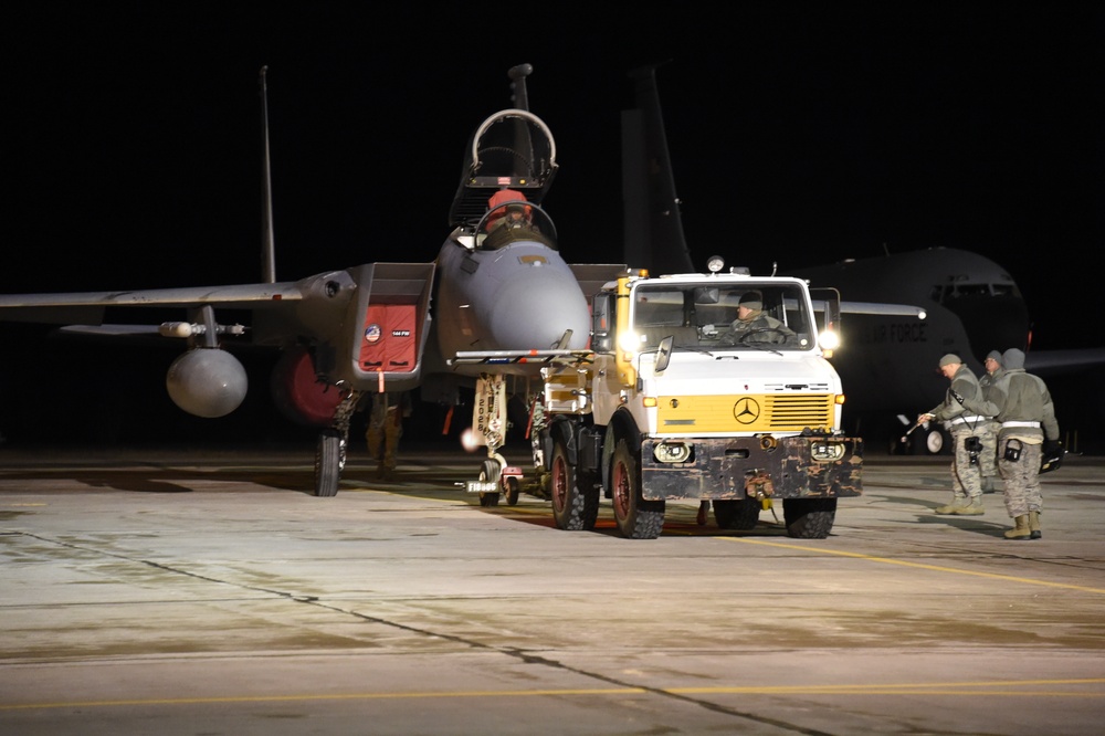 A nighttime military flight line operation showing a fighter jet being towed by a Mercedes-Benz Unimog tow vehicle on an airfield tarmac. Ground crew personnel in military uniforms are visible attending to the aircraft. A large transport aircraft is partially visible in the background.