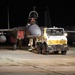 A nighttime military flight line operation showing a fighter jet being towed by a Mercedes-Benz Unimog tow vehicle on an airfield tarmac. Ground crew personnel in military uniforms are visible attending to the aircraft. A large transport aircraft is partially visible in the background.