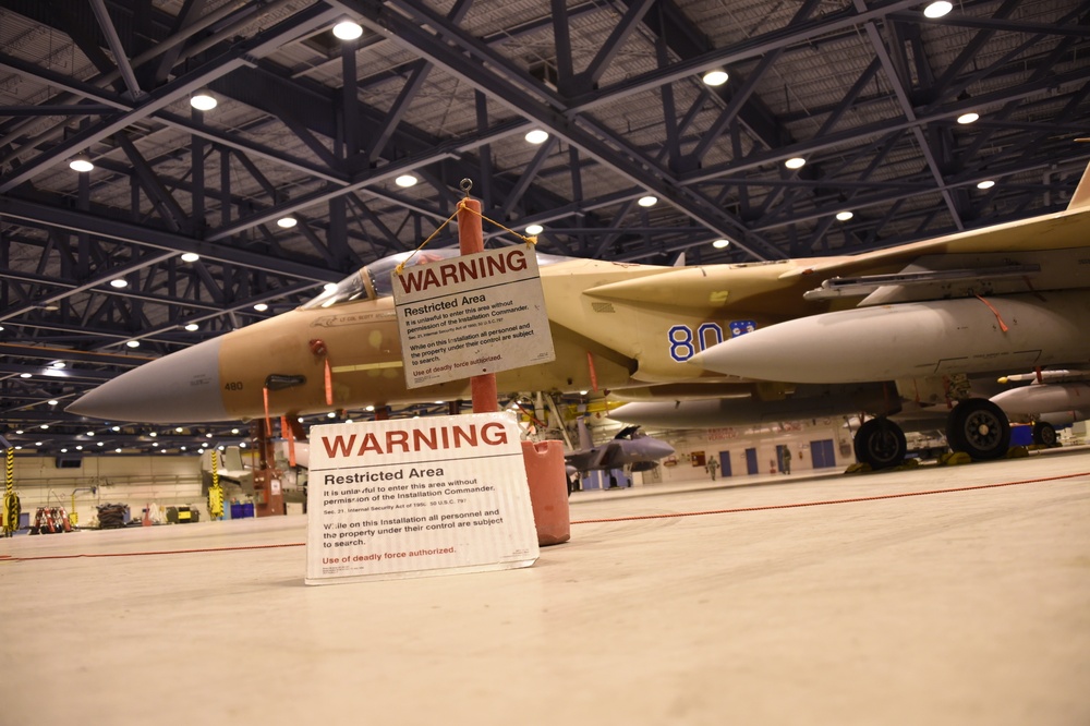 A military jet aircraft with desert tan/sand camouflage paint scheme is parked inside a large hangar facility. Two 'WARNING - Restricted Area' signs are prominently displayed in the foreground, indicating this is a secured military installation. The aircraft bears the number '80' on its fuselage and appears to be an F-15 Eagle variant.