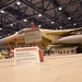 A military jet aircraft with desert tan/sand camouflage paint scheme is parked inside a large hangar facility. Two 'WARNING - Restricted Area' signs are prominently displayed in the foreground, indicating this is a secured military installation. The aircraft bears the number '80' on its fuselage and appears to be an F-15 Eagle variant.
