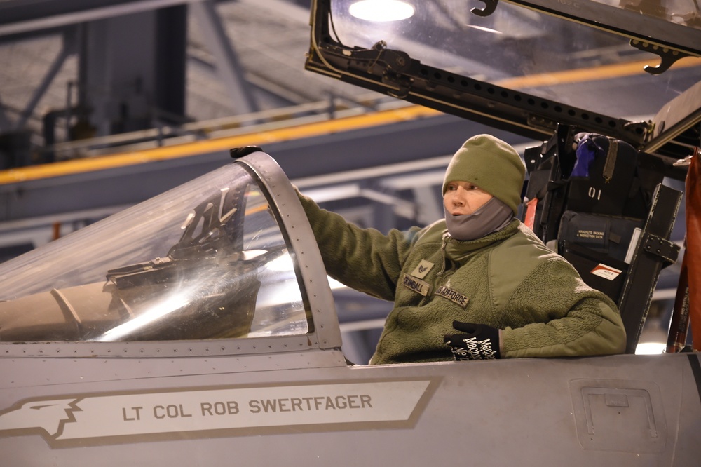 A U.S. Air Force personnel member in cold-weather gear is inspecting or working on a military fighter jet cockpit inside a hangar. The aircraft canopy is open and the fuselage bears the name 'LT COL ROB SWERTFAGER'. The individual is wearing a green fleece jacket with a name tape reading 'SPINDALL' and a U.S. Air Force patch.