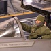 A U.S. Air Force personnel member in cold-weather gear is inspecting or working on a military fighter jet cockpit inside a hangar. The aircraft canopy is open and the fuselage bears the name 'LT COL ROB SWERTFAGER'. The individual is wearing a green fleece jacket with a name tape reading 'SPINDALL' and a U.S. Air Force patch.