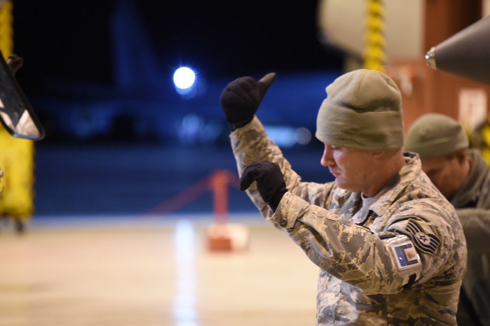 A U.S. Air Force service member in ABU (Airman Battle Uniform) camouflage is photographed at night in what appears to be an aircraft hangar or flight line. The individual is wearing cold-weather gear including a green beanie and black gloves, with one hand raised in a pointing or signaling gesture. A second service member is partially visible in the background.