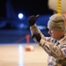 A U.S. Air Force service member in ABU (Airman Battle Uniform) camouflage is photographed at night in what appears to be an aircraft hangar or flight line. The individual is wearing cold-weather gear including a green beanie and black gloves, with one hand raised in a pointing or signaling gesture. A second service member is partially visible in the background.