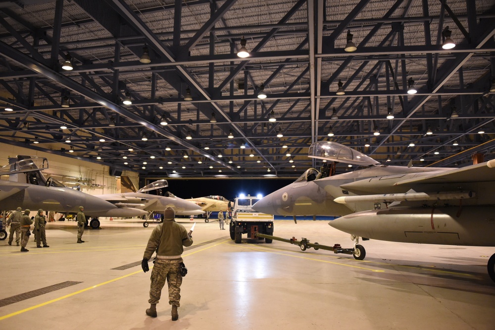 A large military aircraft hangar illuminated by overhead lighting contains multiple fighter jets, likely F-15 Eagles, being serviced or repositioned by ground crew personnel. A tow vehicle is actively moving one of the aircraft while several uniformed personnel are visible throughout the hangar floor. The scene appears to be a nighttime or evening maintenance/repositioning operation at a military air base.
