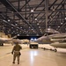 A large military aircraft hangar illuminated by overhead lighting contains multiple fighter jets, likely F-15 Eagles, being serviced or repositioned by ground crew personnel. A tow vehicle is actively moving one of the aircraft while several uniformed personnel are visible throughout the hangar floor. The scene appears to be a nighttime or evening maintenance/repositioning operation at a military air base.