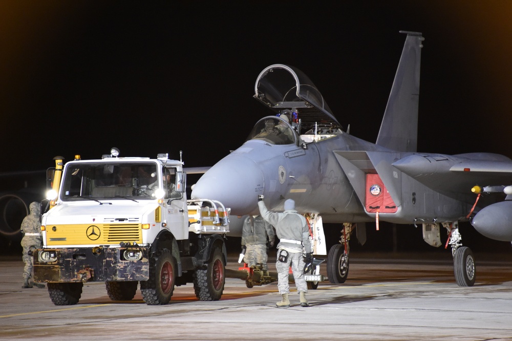 A nighttime military flight line scene showing an F-15 Eagle fighter jet being serviced or prepared for a mission. Ground crew personnel in flight suits are attending to the aircraft alongside a white Mercedes-Benz Unimog support vehicle. The cockpit canopy is open and a pilot appears to be seated in the aircraft.