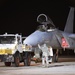 A nighttime military flight line scene showing an F-15 Eagle fighter jet being serviced or prepared for a mission. Ground crew personnel in flight suits are attending to the aircraft alongside a white Mercedes-Benz Unimog support vehicle. The cockpit canopy is open and a pilot appears to be seated in the aircraft.