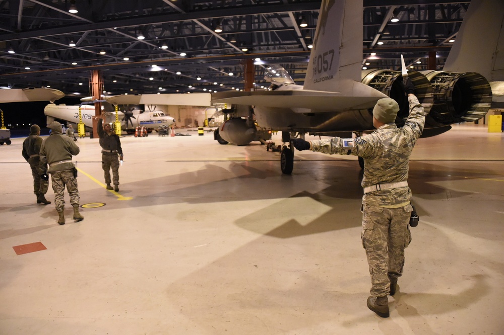 Military personnel are marshaling a fighter jet inside a large aircraft hangar. The aircraft, marked with tail number '057' and the word 'CALIFORNIA,' is being guided by ground crew using hand signals. The hangar is well-lit and contains at least one additional aircraft visible in the background.