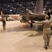 Military personnel are marshaling a fighter jet inside a large aircraft hangar. The aircraft, marked with tail number '057' and the word 'CALIFORNIA,' is being guided by ground crew using hand signals. The hangar is well-lit and contains at least one additional aircraft visible in the background.