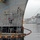 Workers in safety gear are performing maintenance or mooring operations on a large gray naval vessel at a shipyard. A floating work platform/barge with scaffolding is positioned alongside the ship's bow, with personnel at multiple levels. An orange boom or containment barrier is visible floating in the water around the work area, suggesting environmental protection measures.