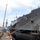 A large gray US Navy vessel bearing the hull number '19' is moored at a naval shipyard facility, likely Yokosuka Naval Base in Japan based on the surrounding infrastructure. The ship is secured with multiple mooring lines in blue and orange, and workers in safety gear are visible on a floating work platform alongside the hull. An American flag is visible at the ship's stern, and various industrial cranes and port equipment surround the scene.