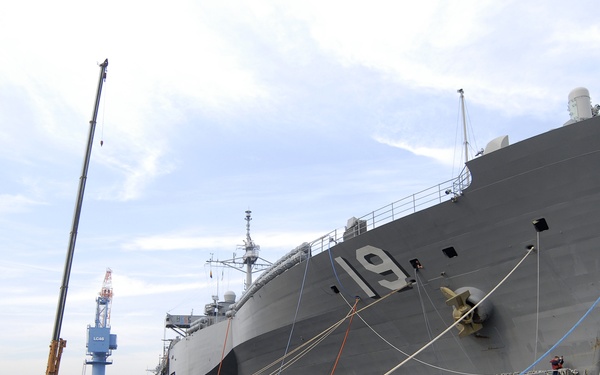 A large gray US Navy vessel bearing the hull number '19' is moored at a naval shipyard facility, likely Yokosuka Naval Base in Japan based on the surrounding infrastructure. The ship is secured with multiple mooring lines in blue and orange, and workers in safety gear are visible on a floating work platform alongside the hull. An American flag is visible at the ship's stern, and various industrial cranes and port equipment surround the scene.
