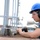 A worker wearing personal protective equipment (PPE) is operating a power drill or pneumatic tool on a metal surface in an industrial or construction setting. The individual is focused on the task, directing the tool toward a metal fixture or post base. The environment appears to be an outdoor industrial worksite, possibly a ship deck or heavy construction platform.