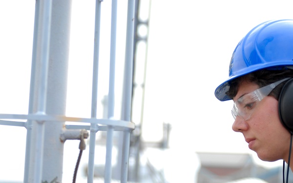 A worker wearing personal protective equipment (PPE) is operating a power drill or pneumatic tool on a metal surface in an industrial or construction setting. The individual is focused on the task, directing the tool toward a metal fixture or post base. The environment appears to be an outdoor industrial worksite, possibly a ship deck or heavy construction platform.