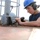 A worker wearing a blue hard hat, safety glasses, and hearing protection is using a power tool on a metal surface, likely aboard a naval vessel or industrial platform. The individual appears to be performing maintenance or repair work on a metal post or fitting secured to a deck surface. The setting suggests an outdoor industrial or maritime environment.