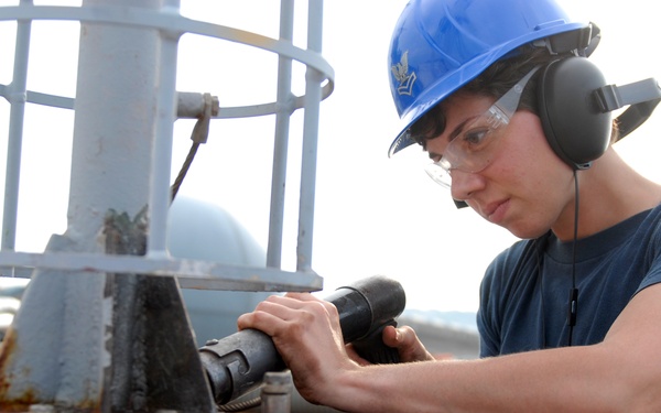 A worker wearing a blue hard hat, safety glasses, and hearing protection is using a power tool on a metal surface, likely aboard a naval vessel or industrial platform. The individual appears to be performing maintenance or repair work on a metal post or fitting secured to a deck surface. The setting suggests an outdoor industrial or maritime environment.