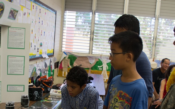 A U.S. Army soldier wearing a lei and IX Corps patch is interacting with students at what appears to be a school recruitment or educational display event. Two young students are examining an 'Army Strong' display table containing printed materials, cards, and promotional items. The setting appears to be a school classroom or gymnasium decorated with student projects and bulletin boards.