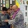 A U.S. Army soldier wearing digital camouflage (ACU) uniform and a purple/white lei is helping a young boy try on a tactical vest at what appears to be a school or community outreach event. The display board behind them shows U.S. Army Public Affairs materials, suggesting this is a military recruitment or educational demonstration. Several children are observing the interaction in what appears to be a school cafeteria or gymnasium.