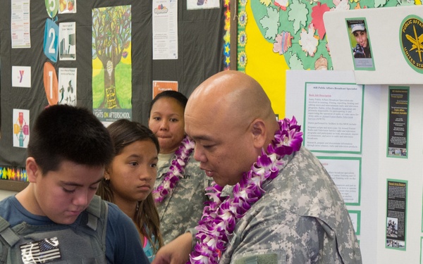 A U.S. Army soldier wearing digital camouflage (ACU) uniform and a purple/white lei is helping a young boy try on a tactical vest at what appears to be a school or community outreach event. The display board behind them shows U.S. Army Public Affairs materials, suggesting this is a military recruitment or educational demonstration. Several children are observing the interaction in what appears to be a school cafeteria or gymnasium.