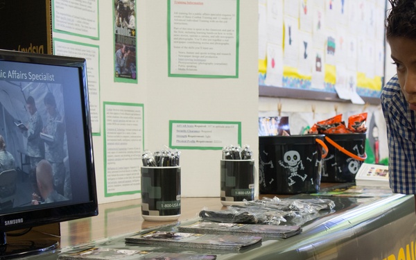 A young boy is leaning over a U.S. Army recruitment display table, examining the materials. The table features a Samsung monitor displaying 'Army MOS 46Q Public Affairs Specialist,' along with brochures, pens, and Halloween-themed candy buckets. The setting appears to be a school or community event with educational posters visible in the background.