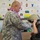 A U.S. Army soldier wearing ACU (Army Combat Uniform) and a floral lei is helping a child try on a military helmet in what appears to be a classroom setting. The background displays colorful student artwork and paper projects pinned to a bulletin board. This appears to be a school visit or educational outreach event by military personnel.