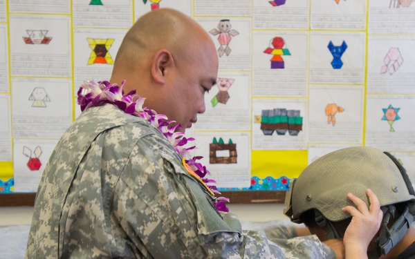 A U.S. Army soldier wearing ACU (Army Combat Uniform) and a floral lei is helping a child try on a military helmet in what appears to be a classroom setting. The background displays colorful student artwork and paper projects pinned to a bulletin board. This appears to be a school visit or educational outreach event by military personnel.