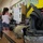 A classroom setting appears to be hosting a military equipment display or educational event, with children gathered in the background examining items on tables. In the foreground, military gear including a tactical helmet, body armor vest, and digital camouflage equipment are displayed on a wooden ledge. The setting appears to be an elementary school based on the colorful bulletin boards and classroom decor visible on the walls.