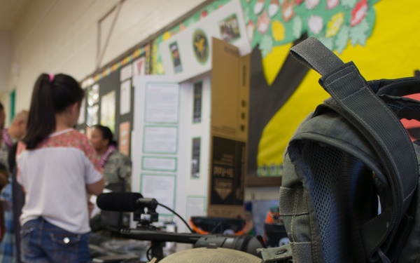 A classroom setting appears to be hosting a military equipment display or educational event, with children gathered in the background examining items on tables. In the foreground, military gear including a tactical helmet, body armor vest, and digital camouflage equipment are displayed on a wooden ledge. The setting appears to be an elementary school based on the colorful bulletin boards and classroom decor visible on the walls.