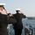 Two naval officers in dress uniforms are standing on the deck of a warship, rendering a formal salute toward a large vessel passing in the background. The officers are viewed from behind, wearing white-topped naval caps and dark dress jackets with gold sleeve stripes indicating senior rank. This appears to be a formal naval courtesy salute, a traditional maritime protocol when ships pass one another.