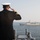 A naval officer in formal dress uniform is standing on the deck of a warship, rendering a salute toward a passing naval vessel in open water. The officer's back is to the camera, displaying gold sleeve stripes indicative of senior rank. The scene appears to depict a formal naval courtesy exchange between two ships at sea.