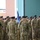 A military formation ceremony is taking place, likely indoors or under a covered structure. Soldiers wearing US Army OCP (Operational Camouflage Pattern) uniforms and black berets are standing in organized ranks. Color guard members are visible holding guidons/flags on the left side of the formation.