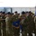 A military ceremony is taking place, likely a casing or uncasing of colors, where two soldiers in the foreground are folding or handling a blue unit guidon/flag with gold fringe. Multiple soldiers in US Army MultiCam (OCP) uniforms and black berets are standing in formation in the background. The setting appears to be a military installation, possibly a hangar or outdoor staging area.