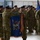 A military ceremony is taking place indoors, likely in a hangar or large facility, where two soldiers in the foreground are handling a blue unit guidon or organizational color with gold fringe and campaign streamers. The flag displays an eagle and other military insignia on a blue field. Rows of soldiers in Army Combat Uniforms (ACUs/OCP pattern) and black berets stand in formation in the background.