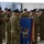 A military ceremony is taking place indoors, likely a casing or uncasing of colors ceremony. Two soldiers in the foreground are handling a blue unit guidon/flag adorned with multiple campaign streamers and a finial on the staff. A formation of soldiers in camouflage uniforms and black berets stands at attention in the background.