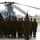A formation of military personnel in camouflage uniforms and black berets stand at attention on a tarmac or airfield apron. Behind them is a large tandem-rotor helicopter, identifiable as a CH-47 Chinook. A blue military unit guidon flag is visible in the background, suggesting a formal military ceremony or unit formation.