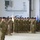 A large formation of military personnel in camouflage uniforms and black berets are standing at attention inside what appears to be a military hangar or large indoor facility. Two unit guidons are visible — one white and one blue — being carried by soldiers within the formation. The scene is consistent with a formal military ceremony such as a change of command, activation, or unit formation.