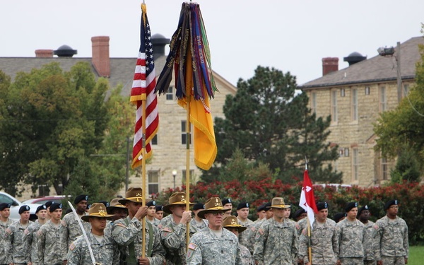 A U.S. Army military ceremony is taking place on a parade field, likely at a stateside installation. Soldiers in Army Combat Uniforms (ACU) are assembled in formation, with a color guard carrying the U.S. national flag and a unit guidon with campaign streamers. The background features historic stone and brick buildings consistent with an established military post.