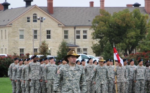 A military formation ceremony is taking place on a green parade field in front of a large stone institutional building. A senior non-commissioned officer wearing a distinctive campaign (drill sergeant) hat stands at the front of the formation, facing away from the assembled troops. Multiple soldiers in ACU (Army Combat Uniform) pattern camouflage are standing at attention in organized ranks behind him.