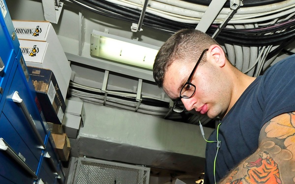 A young man with glasses and a prominent full-sleeve tattoo featuring a tiger design is working in what appears to be a storage or supply room, sorting through boxes and small parts. The environment has blue metal shelving units, overhead cable bundles, and a metal mesh cage door visible in the background, suggesting a military or industrial setting. The angle is a low, upward-looking perspective that emphasizes the tattoo and the cluttered workspace.