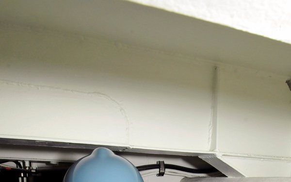 A worker wearing a blue hard hat and clear safety glasses peers into a confined space beneath a raised floor or equipment panel. She is dressed in an olive/green long-sleeve shirt and is resting her hands on the edge of the opening while inspecting the area below. The setting appears to be an industrial or technical facility with cabling and equipment visible in the confined space.