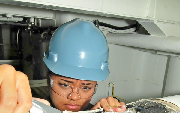 A worker wearing a blue hard hat and safety glasses is using a scraper or chisel tool to remove deteriorated insulation or coating from a large pipe or surface in a confined industrial space. The area being worked on shows significant corrosion, black degraded material, and damaged white insulation. The setting appears to be a mechanical or utility space, possibly aboard a naval vessel or industrial facility.