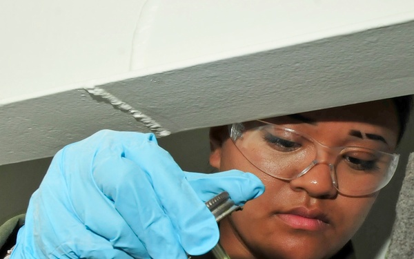 A person wearing safety glasses and blue nitrile gloves is working in a confined or damaged space, using a small tool (appears to be a screwdriver or probe) to examine or work on debris and broken material. The individual is wearing an olive/green work uniform and appears to be conducting inspection, repair, or sample collection work. The surrounding area shows broken concrete, rubble, and structural debris.