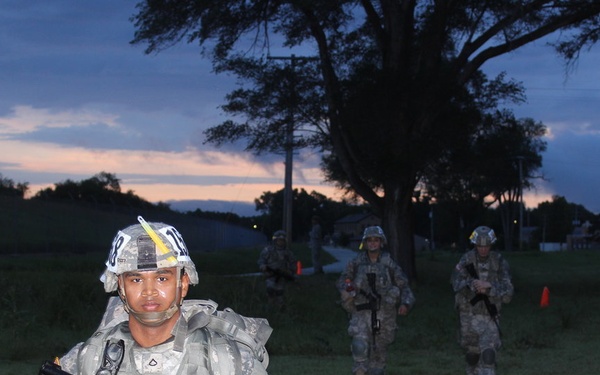 The image shows military personnel in U.S. Army ACU (Army Combat Uniform) conducting what appears to be a training exercise at dusk or dawn. The foreground soldier is carrying a rifle with a training device attachment (blue/orange tip indicating a MILES or Simunition training system) and wearing a helmet with markings. Two additional soldiers are visible in the background on a grassy field.