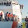 A U.S. Coast Guard vessel is docking at a pier, with crew members in orange survival suits and blue hard hats handling mooring lines along the ship's deck. Personnel on the bridge wings and upper decks observe the docking operation, while dock workers on the pier monitor the process. The scene depicts a coordinated ship-to-shore mooring evolution in what appears to be a harbor or naval station.