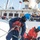 The image shows a group of personnel, likely U.S. Coast Guard members, working on the deck of a vessel handling mooring lines and rigging. The foreground figure wears a blue hard hat, navy coveralls, and an orange life vest while gripping a thick rope along a metal railing. Additional crew members in similar gear are visible in the background, along with observers on an upper deck.