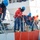 A crew of maritime personnel wearing orange life vests, blue helmets, and navy uniforms are working together on the deck of a white vessel, handling mooring lines. The scene appears to depict a docking or undocking operation, with multiple crew members pulling and managing thick ropes secured to cleats and bollards. Additional personnel are visible on an upper deck in the background observing or supervising the operation.