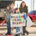 A group of family members, including two women and two children, are gathered outdoors holding a handmade 'Welcome Home Daddy!' sign decorated with colorful letters, stars, and blue pom-poms. The scene appears to be a military or deployment homecoming event, likely at a base or airfield. The participants are dressed in winter clothing and appear joyful and excited.