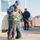 A U.S. Coast Guard service member in uniform is being embraced by three children in what appears to be a homecoming or reunion scene at a military facility. Two other Coast Guard personnel stand in the background observing the emotional moment. The setting appears to be an outdoor dock or base area with shipping crates visible in the background.